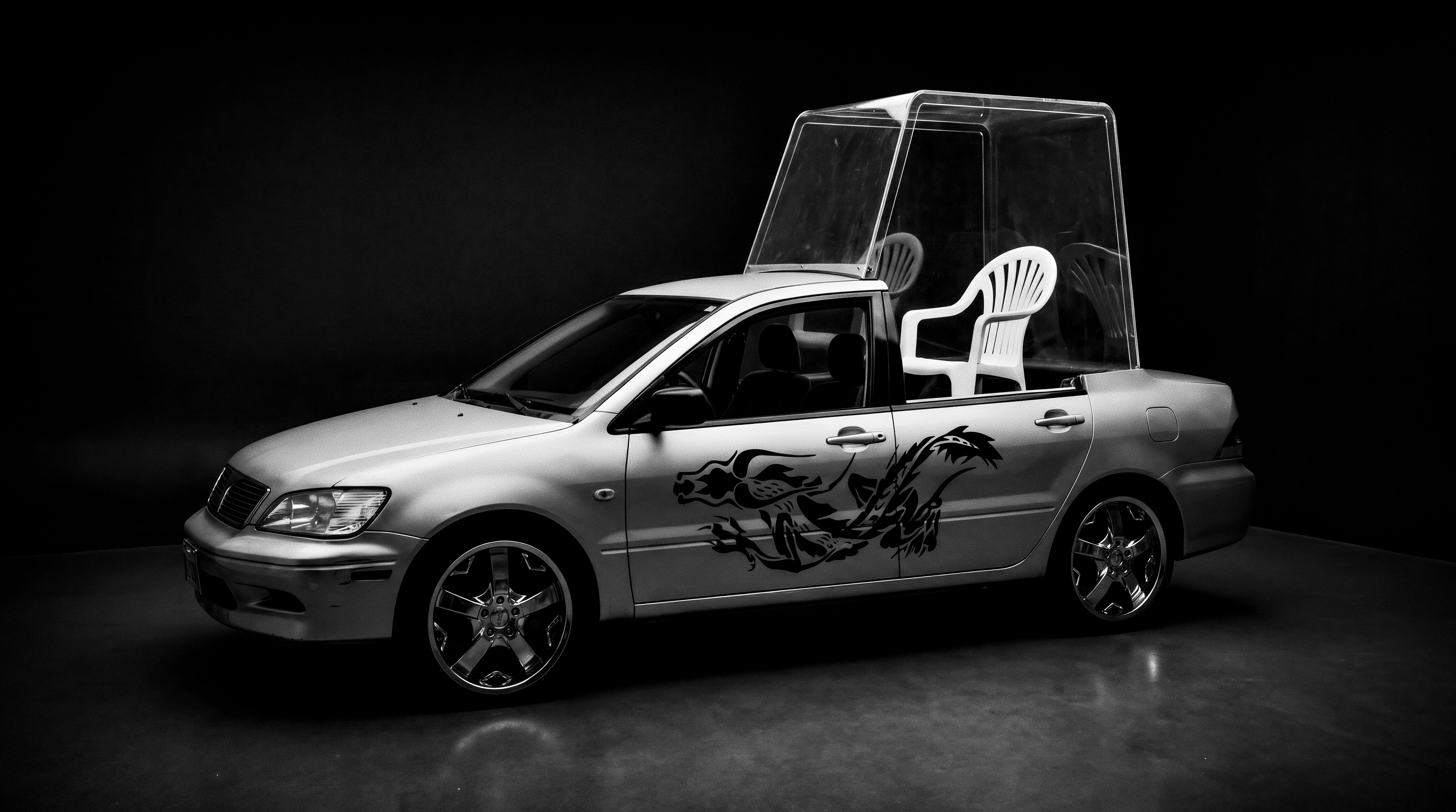 Black-and-white image of a silver car with a dragon graphic and a white plastic chair inside a tall transparent enclosure.