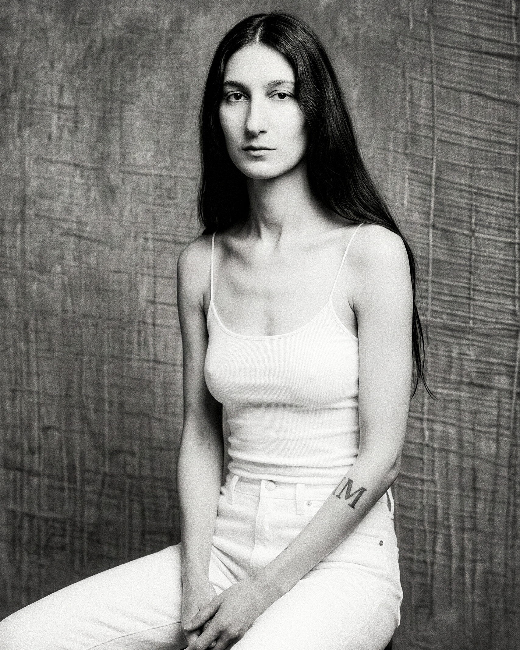 Black-and-white portrait of a seated figure in a white tank against a textured backdrop.