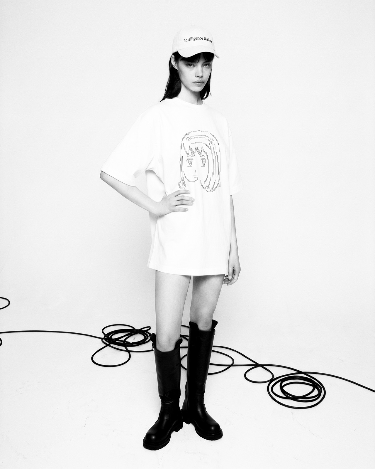 Recent Work — a black-and-white studio full-length: an Intelligence Matters cap and an oversized white tee carrying a face drawn in ASCII characters, black knee boots, studio cable coiled across the floor