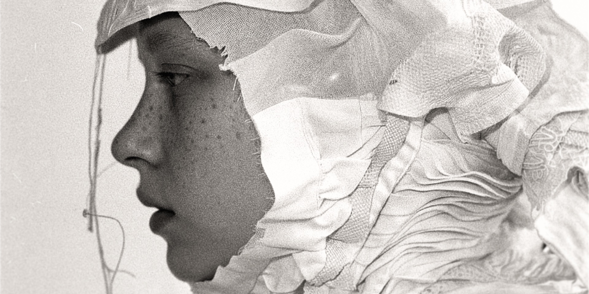 Black-and-white profile portrait — a freckled face framed in a raw-edged patchwork textile hood, pleated fabric at the shoulder, on film grain.