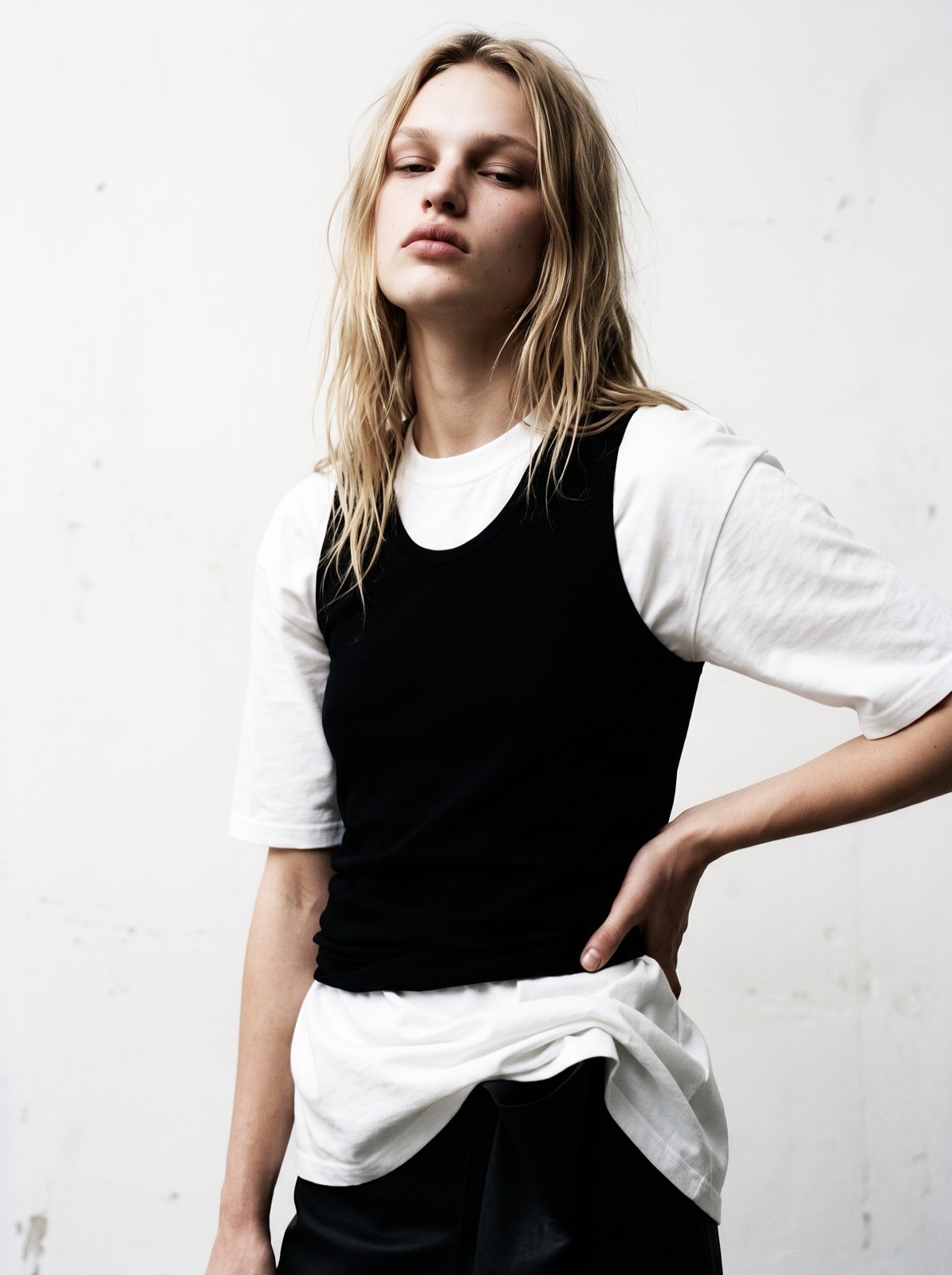 Image — a lookbook frame against a scuffed white wall: a black tank layered over a white tee, hand on the hip