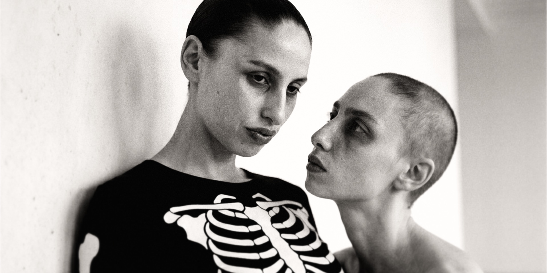 Negative Space — two figures against a white wall: one in a skeleton-print top reading down, one shaved-head in profile reading up, black-and-white.