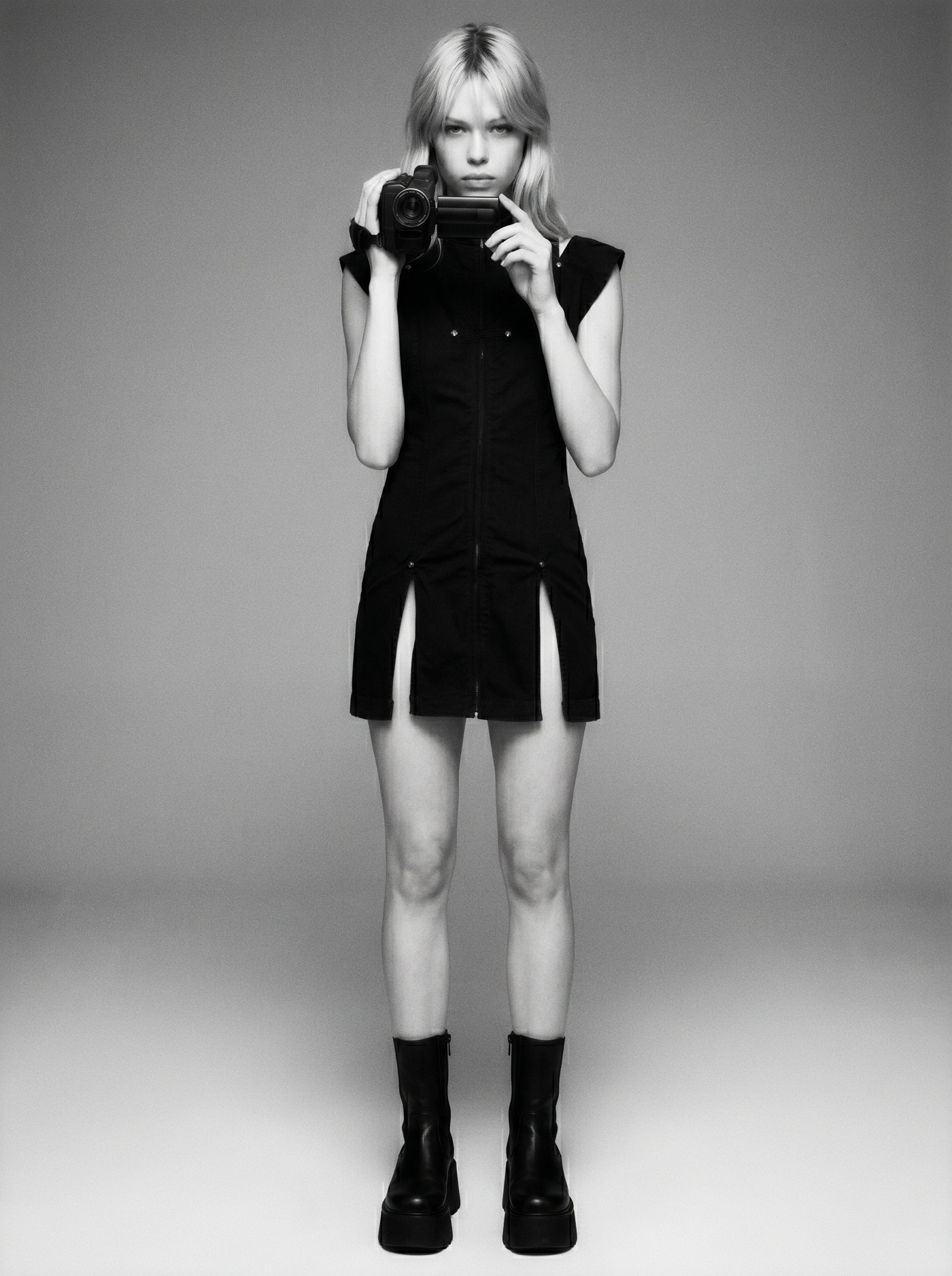 Image Production — a black-and-white studio full-length: a figure in a black zip-front dress and platform boots raising a camcorder back at the lens, on a grey sweep