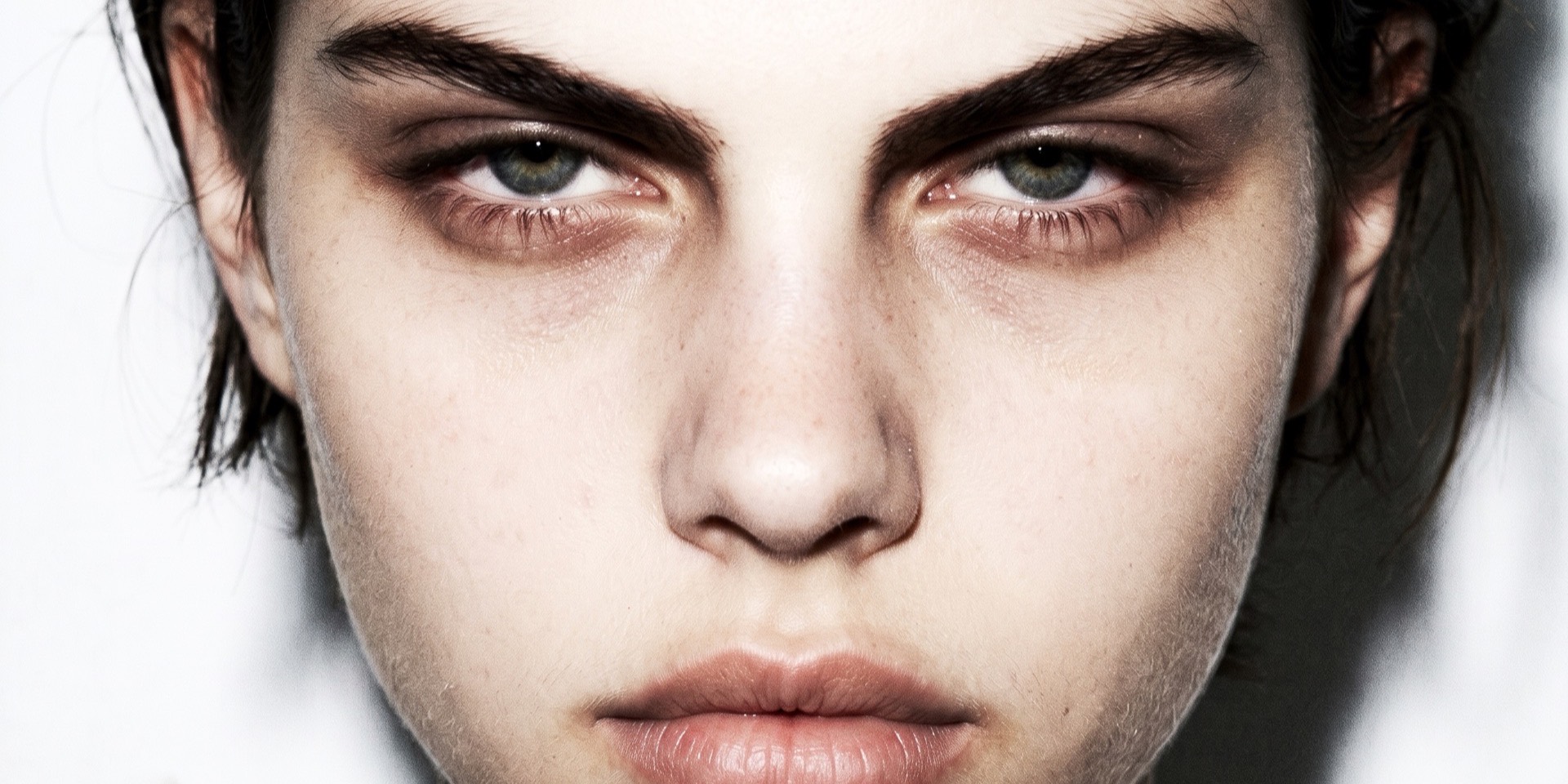 Extreme frontal close-up — the face filling the frame, dark brows, grey-green eyes level with the lens, skin left unretouched against a near-white ground.