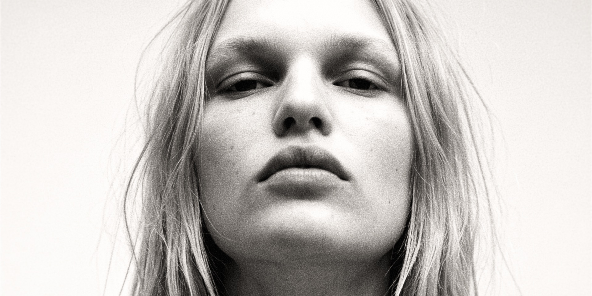 Black-and-white frontal close-up — chin lifted, eyes reading down into the lens, loose hair, on film grain.
