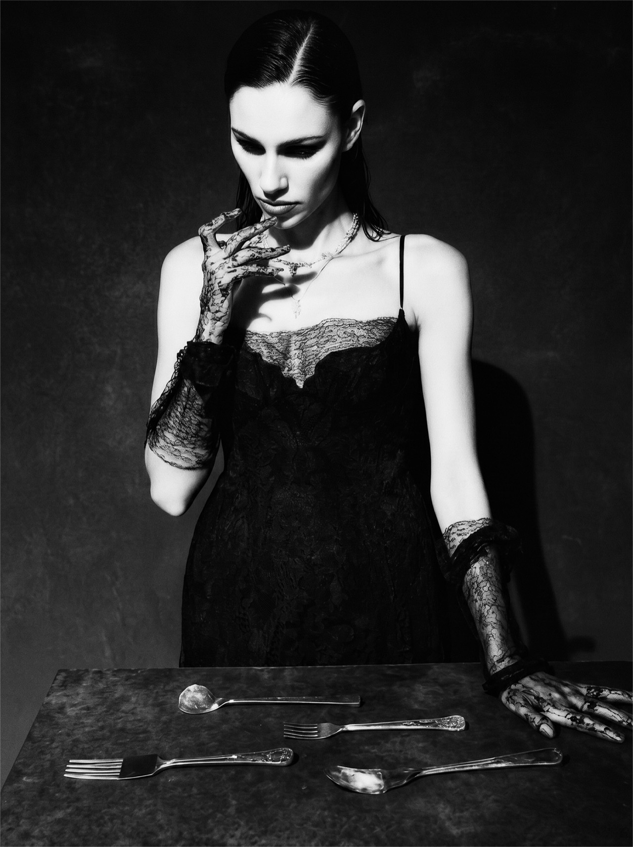 NIIA — gothic chiaroscuro portrait: black slip, skeletal-lace gloves at a dark table set with scattered silverware, downcast and withheld.