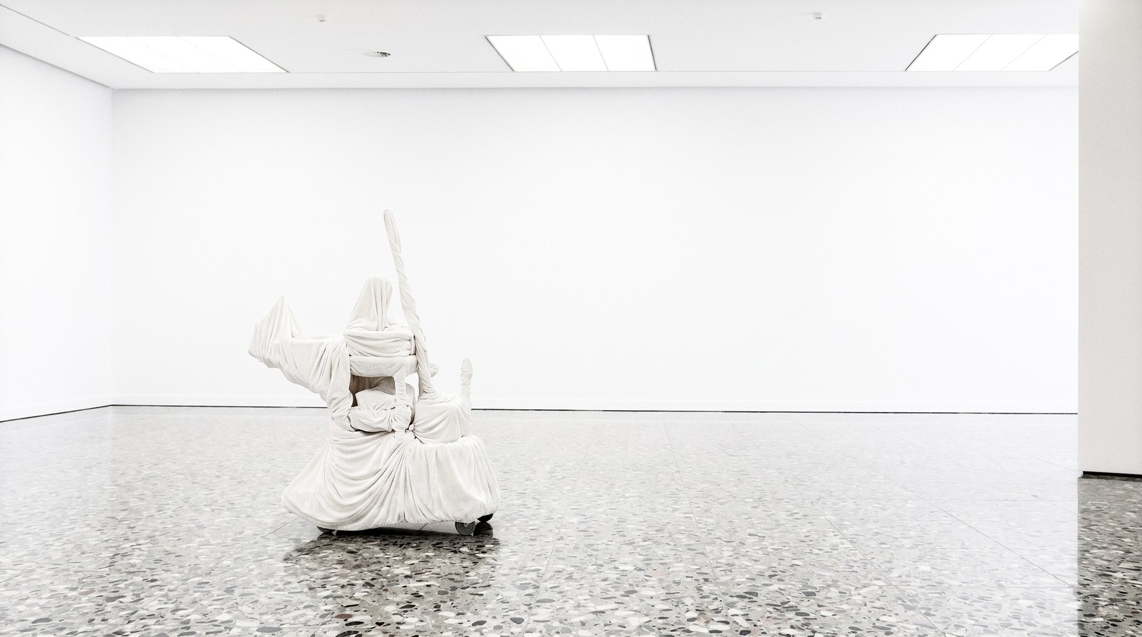 An empty white gallery with a terrazzo floor and a single white-shrouded form on castors standing mid-room.